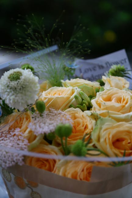 A close-up of a floral arrangement featuring peach-colored roses with delicate petals, interspersed with white pom-pom-like flowers and small green buds. The bouquet is wrapped in light-colored paper 