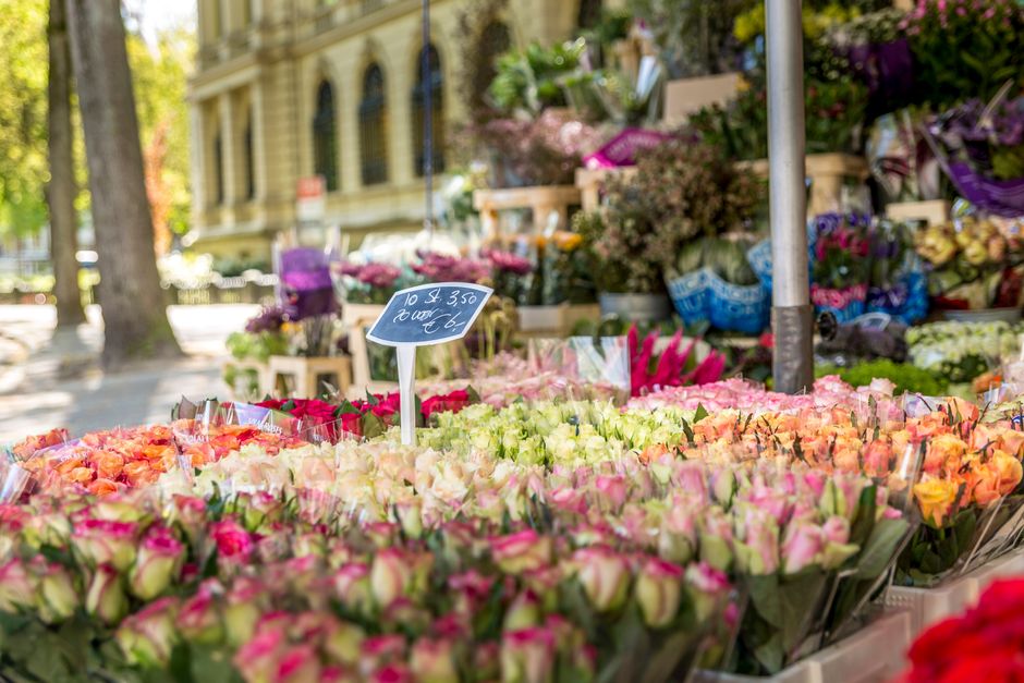 A vibrant outdoor flower market display featuring numerous fresh bouquets and individual flower arrangements. The foreground showcases a variety of roses in shades of pink, peach, and cream, tightly p