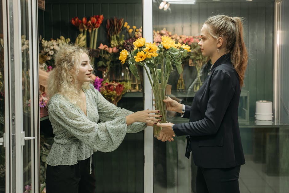 A floral arrangement is being handed over by a woman with curly blonde hair, dressed in a pale green, long-sleeved blouse, to a female florist with long, light brown hair tied back, wearing a dark bla