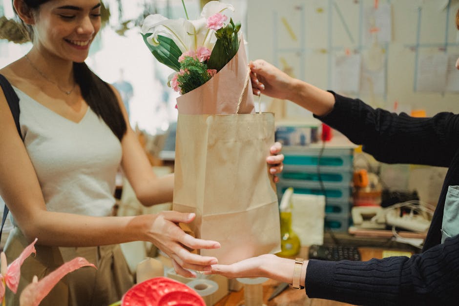 A woman in a white sleeveless top is smiling as she receives a bouquet of fresh flowers, including white lilies and pink roses, wrapped in pale pink tissue paper and brown paper from a florist in a bl