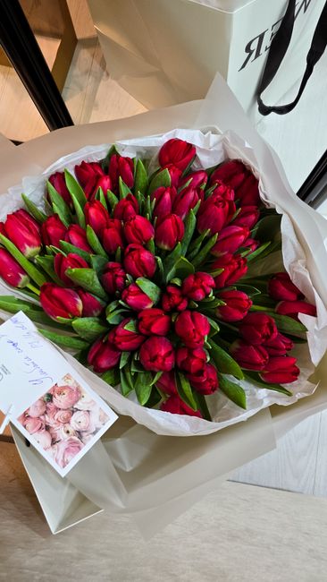 A large bouquet of fresh red tulips with green stems and leaves, wrapped in white paper with a black ribbon, resting on a surface. The tulips are tightly packed and appear vibrant, with smooth petals