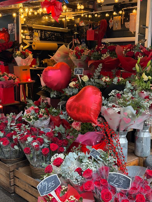 A vibrant display of floral arrangements and gifts at a flower shop, featuring clusters of red roses, white baby's breath, and greenery. Two heart-shaped helium balloons in glossy red float above the 
