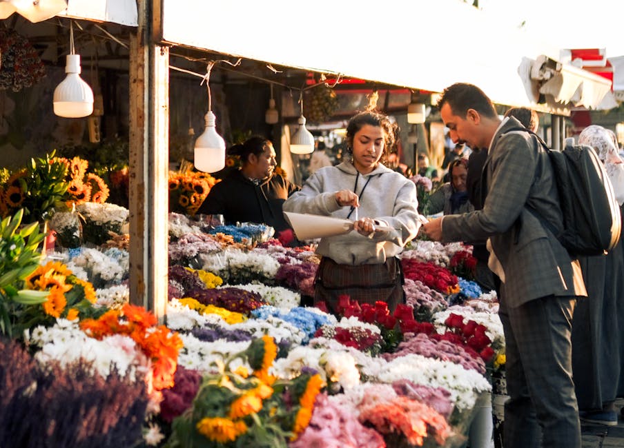 A flower stall at an outdoor market displays a vibrant array of fresh flowers, including sunflowers, chrysanthemums, roses, and daisies in shades of yellow, white, pink, red, purple, and orange, arran